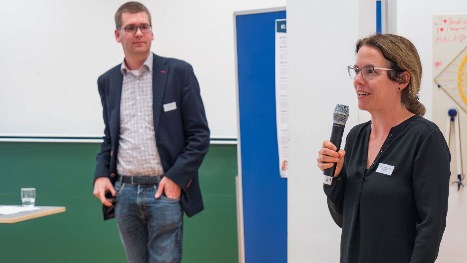 Eine Frau hält ein Mikrofon und steht vor einer Pinnwand. Im Hintergrund steht ein Mann
